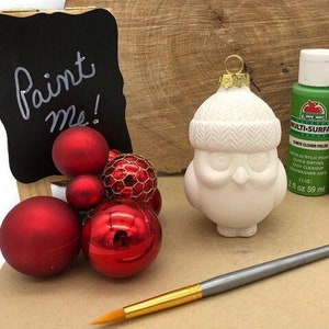 May include: A white ceramic owl ornament with a knitted hat, surrounded by red Christmas ornaments, green and red acrylic paints, and a paintbrush. A small chalkboard sign reads "Paint Me!".