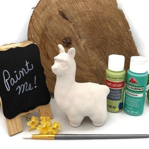 Puede incluir: Figura de llama de cerámica blanca con un cartel que dice "¡Píntame!" y dos botellas de pintura acrílica, una verde y una azul, sobre una superficie de madera.
