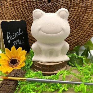 Könnte beinhalten: Eine weiße Keramik-Froschfigur mit einem lächelnden Gesicht, die auf einem Holzsockel sitzt. Der Frosch ist bereit zum Bemalen. Das Schild sagt "Bemal mich!"
