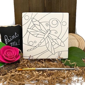 May include: A white ceramic tile with a black outline of a dragonfly and a swirling pattern. The tile is on a stand and has a small sign that says "Paint Me!"