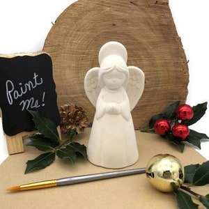 Könnte beinhalten: Weiße Keramikengelfigur mit Flügeln und Heiligenschein. Der Engel hält seine Hände zum Gebet zusammen. Die Figur steht auf einer Holzoberfläche mit einem Schild, auf dem "Bemal mich!" steht, und etwas Grün.