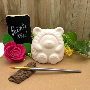 Può includere: Figurina di orso in ceramica bianca con un cartello "dipingimi". L'orso è seduto con le zampe davanti a sé. Incluso un pennello.