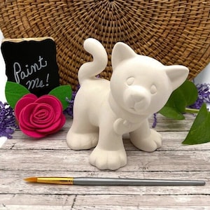May include: White ceramic cat figurine with a paint me sign. The cat is standing on a wooden surface with a paintbrush in front of it.