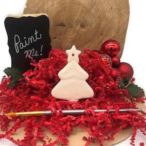 K&ouml;nnte beinhalten: Wei&szlig;er Keramik-Weihnachtsbaum-Anh&auml;nger mit einem Stern oben. Der Anh&auml;nger ist von rotem zerkleinertem Papier und einem Pinsel umgeben. Ein schwarzes Schild aus Schiefertafel sagt "Bemal mich!"