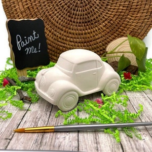May include: A white ceramic car figurine with rounded features sits on a wooden surface, surrounded by green paper grass. A small chalkboard sign that says "Paint Me!" is in the background. A paintbrush is in the foreground.