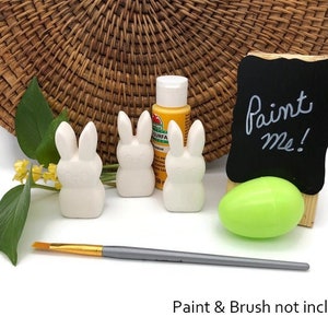 Puede incluir: Tres figuritas de conejo de cerámica blanca con un cartel de "píntame", un huevo verde y una pequeña botella de pintura amarilla. Pintura y pincel no incluidos.