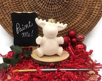 Peut inclure: Ornement de renne en céramique blanche avec un dessus doré, prêt à être peint. Il est posé sur une base en bois avec du papier déchiqueté rouge et un petit tableau noir qui dit "Peignez-moi !"