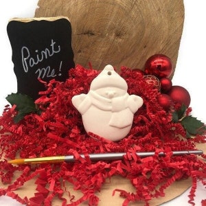 May include: White ceramic snowman ornament with a paint me sign. The ornament is surrounded by red shredded paper and red and gold Christmas ornaments.