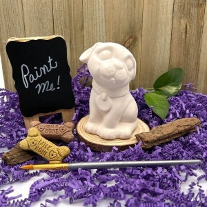 May include: White ceramic dog figurine with a collar, sitting on a wooden base. The figurine is surrounded by purple shredded paper and a paintbrush. A chalkboard sign reads "Paint Me!"
