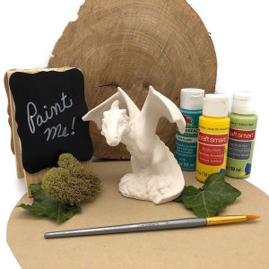 Puede incluir: Figurilla de dragón de cerámica blanca con las alas extendidas, lista para ser pintada. El dragón está sentado sobre una base y está acompañado de un pincel, un pequeño letrero de pizarra que dice "¡Píntame!" y tres botellas de pintura acrílica en amarillo, verde y azul.