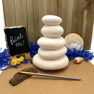 May include: A white ceramic stack of stones, ready to be painted. The stack is on a brown surface with a paintbrush and a small wooden sign that says "Paint me!"
