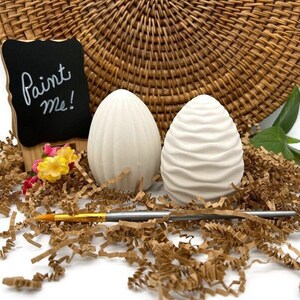 May include: Two white ceramic eggs, one smooth and one with a textured surface, are sitting on brown shredded paper. A paintbrush is lying on the paper. A small chalkboard sign says "Paint Me!"