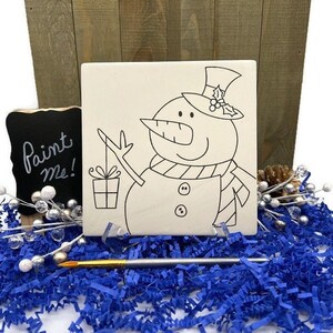 May include: A white ceramic tile with a black line drawing of a snowman holding a gift. The tile is ready to be painted. The words "Paint Me!" are written on a black chalkboard in the background.