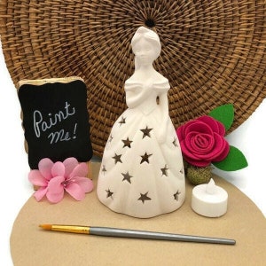 Puede incluir: Figurilla de cerámica blanca de una niña con un vestido con estrellas. La figurita es un proyecto de manualidades para pintar. La imagen también incluye un pequeño letrero de pizarra que dice "¡Píntame!" y una flor rosa.