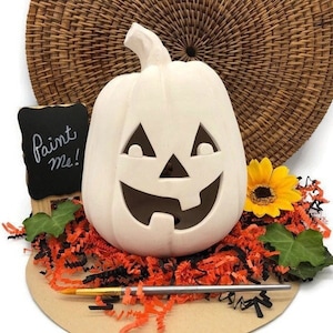 May include: White ceramic pumpkin with a carved jack-o'-lantern face. The pumpkin is sitting on a bed of orange and black shredded paper. A paintbrush and a small chalkboard with the words "Paint me!" are also in the image.