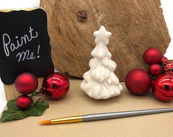 May include: White ceramic Christmas tree ornament with a star on top. The ornament is surrounded by red baubles and a wooden sign that says "Paint Me!" A paintbrush is also included.