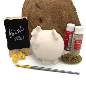 May include: A white ceramic pig piggy bank with a paint me sign, two bottles of acrylic paint, a paintbrush, and a small pile of green moss.