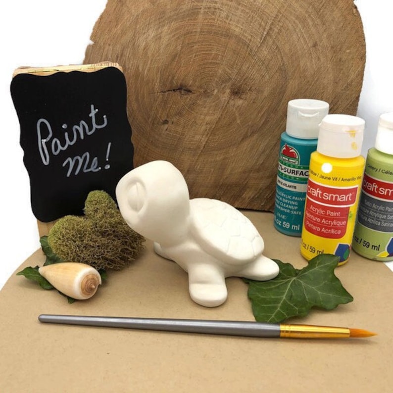 May include: A white ceramic turtle figurine with a paint me sign, paintbrushes, and three bottles of acrylic paint in blue, yellow, and green.