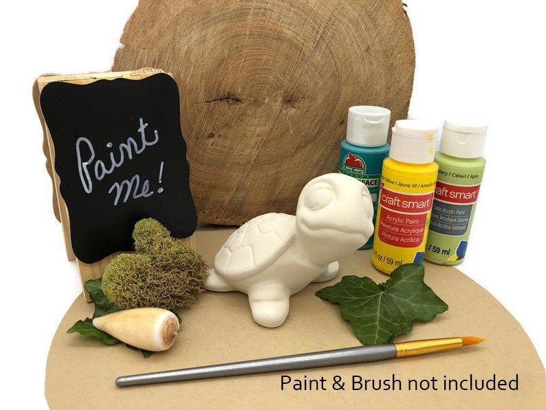 May include: White ceramic turtle figurine with a paint me sign. The turtle is sitting on a wooden surface with a small shell and green moss. Three bottles of acrylic paint are visible in the background.