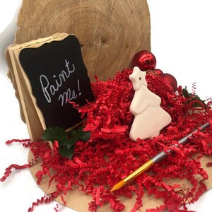 K&ouml;nnte beinhalten: Ein wei&szlig;er Keramik-Weihnachtsbaum-Anh&auml;nger mit einem Stern oben, umgeben von roten Papierschnitzeln. Im Hintergrund befindet sich ein kleines Holz-Staffelei mit einem Schild aus schwarzem Brett, auf dem "Malt mich!" steht. Ein Pinsel ist ebenfalls enthalten.