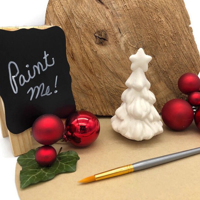 May include: White ceramic Christmas tree with a star on top, surrounded by red ornaments and a paintbrush. A sign reads "Paint Me!"