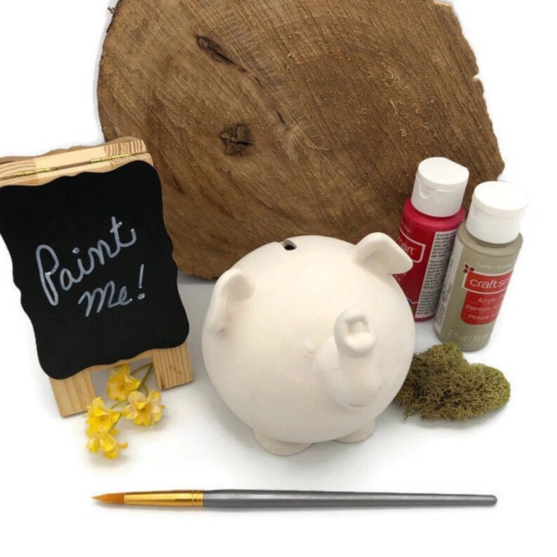 May include: White ceramic piggy bank with a paint me sign, two bottles of acrylic paint, a paintbrush, and yellow flowers.