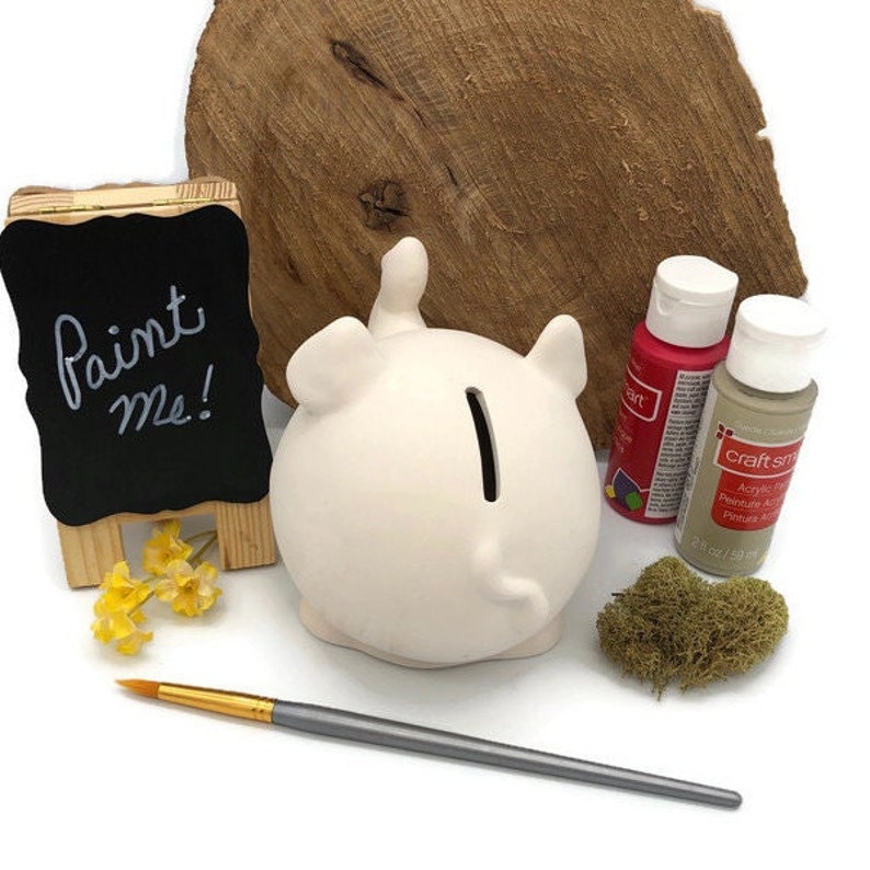 May include: A white ceramic elephant piggy bank with a paint me sign, two bottles of acrylic paint, a paintbrush, and a piece of green moss.