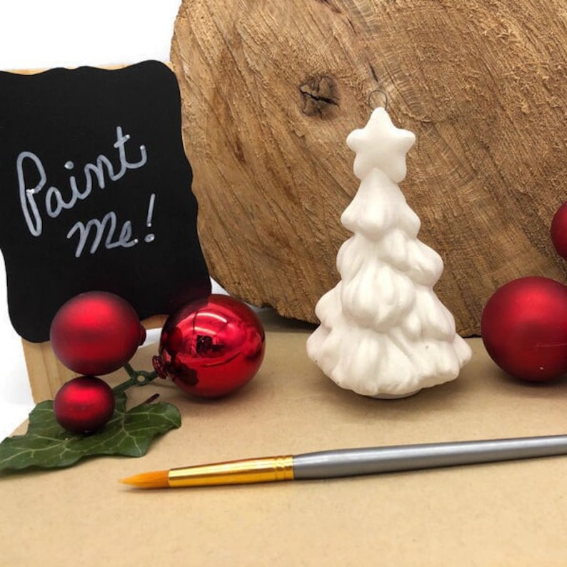 May include: White ceramic Christmas tree ornament with a star on top. The ornament is sitting on a wooden surface with red ornaments and a paintbrush. The sign says "Paint Me!"