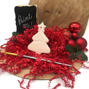 K&ouml;nnte beinhalten: Ein wei&szlig;er Keramik-Weihnachtsbaum-Anh&auml;nger mit einem Stern oben, umgeben von rotem zerkleinertem Papier und einem Pinsel. Der Anh&auml;nger hat die Worte "Paint Me!" auf einer schwarzen Tafel dahinter geschrieben.