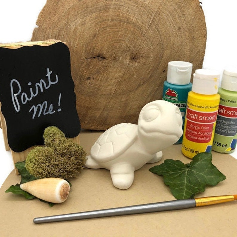 May include: A white ceramic turtle figurine with a paint me sign, paintbrushes, and acrylic paint in blue, yellow, and green.