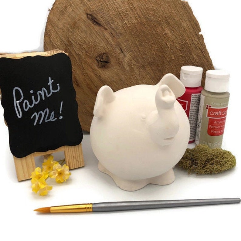 May include: A white ceramic pig figurine with a paint me sign, paintbrushes, and two bottles of acrylic paint.