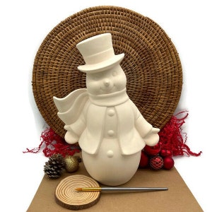 Może przedstawiać: Biała ceramiczna figurka bałwana z czarnym kapeluszem, szalikiem i zapinaną na guziki kurtką. Bałwan siedzi na brązowej powierzchni z czerwonymi i złotymi elementami dekoracyjnymi.
