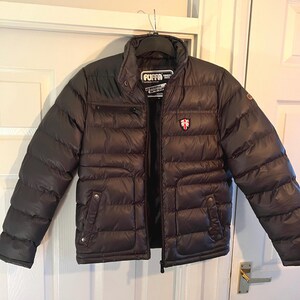 May include: A black puffer jacket with a zipper closure and a logo that says "PUFFA" on the left chest. The jacket has a quilted design and a stand-up collar.
