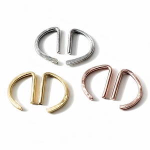 May include: Three pairs of metal earrings in silver, gold, and rose gold. Each pair features a curved, D-shaped design with a straight bar. The earrings are displayed on a white background, showcasing their different finishes and textures.