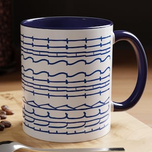 Op de afbeelding: Witte keramische mok met een blauw interieur en handvat. De mok heeft een herhalend blauw lijnenontwerp. Een zilveren lepel en een macaron liggen op een houten oppervlak naast de mok.