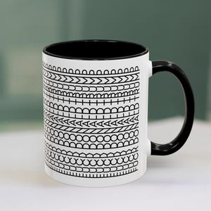May include: A white ceramic mug with a black interior and a black handle. The exterior features a repeating black graphic pattern consisting of horizontal bands of geometric shapes, including circles, triangles, chevrons, and lines.