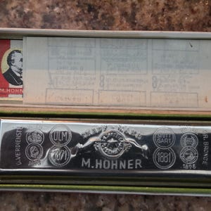 May include: An open, vintage green box containing a silver harmonica. The harmonica is engraved with the words "M. Hohner" and various city names and dates. A small red and white label with a portrait is visible.