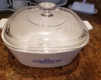 May include: White casserole dish with a clear glass lid and a clear glass knob. The dish has a square shape with rounded corners and white handles. The dish has a blue floral design on the front.