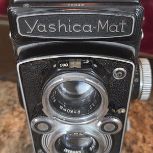 May include: A black Yashica-Mat camera with silver lenses and dials. The camera has the name "Yashica-Mat" printed on the front. The lens is f=80mm, and the camera has a Lumaxar lens.