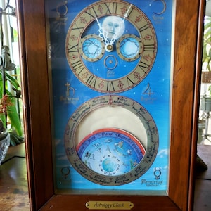 May include: A wooden-framed astrology clock with a blue background. The clock face features Roman numerals, celestial symbols, and a depiction of the moon. The lower section displays a zodiac wheel. The clock has a small gold plaque that reads "Astrology Clock."