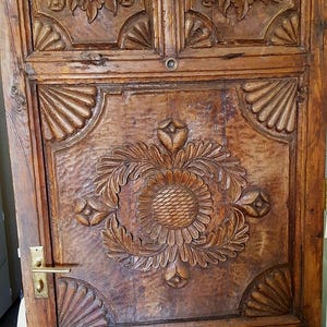 May include: A weathered, wooden door with intricate carvings. The door features a large floral design in the center and decorative panels with heart and shell motifs. The wood has a warm, brown tone with visible grain and a brass handle.
