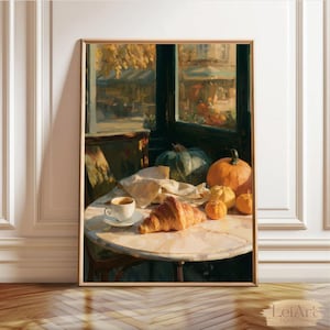 Op de afbeelding: Een ingelijste kunstprint met een cafétafel met een wit koffiekopje, een goudbruine croissant op een schoteltje en verschillende oranje pompoenen. De scène bevindt zich bij een raam met uitzicht op een straatbeeld met herfstkleuren.