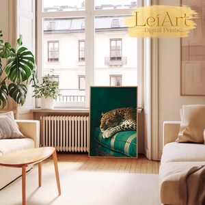 May include: A framed art print featuring a leopard resting on a green and white striped cushion. The leopard has a spotted coat and is set against a dark green background. The print is displayed in a bright, airy room.