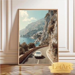 May include: Framed print of a coastal road with a white car driving along the winding asphalt. The image features a scenic view of the ocean, cliffs, and a small town. The print is in a gold frame and is leaning against a white wall.