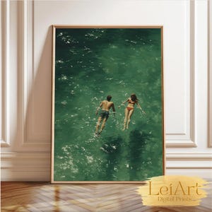May include: A framed print featuring two people swimming in emerald green water. The artwork captures a serene moment with a man and a woman floating in the sea. The print is displayed in a wooden frame.
