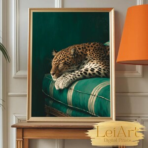 May include: A framed art print featuring a sleeping leopard with a spotted coat, resting on a green striped cushion. The artwork has a gold frame and is set against a dark green background. The print is displayed on a wooden table.