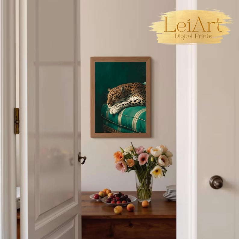 May include: A framed art print featuring a leopard resting on a green striped cushion. The artwork is displayed on a wall, with a vase of flowers and a bowl of fruit on a wooden table in the foreground. The print has the text "LeiArt Digital Prints".