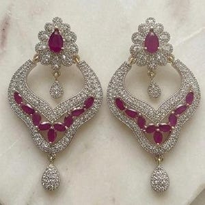 May include: A pair of ornate, gold-toned earrings. Each earring features a cluster of small, clear stones surrounding a large, teardrop-shaped, ruby-colored stone. The earrings have a curved, open design with additional ruby-colored stones and a dangling, teardrop-shaped stone.