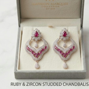 Op de afbeelding: Een paar met robijn en zirkonia bezette chandbali-oorbellen, gepresenteerd in een grijze sieradendoos. De oorbellen hebben een druppelvorm met een bloemmotief en een peervormige steen. De tekst "MANSION MARQUEE COLLECTION EST. 2026" is zichtbaar.