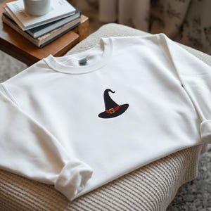 May include: A white sweatshirt featuring a black witch hat with a red band and gold buckle. The long-sleeved top is made of a soft material and is suitable for casual wear. The witch hat design is embroidered on the front.